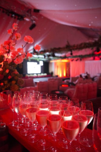 Hurley House - A luxurious event setting with champagne glasses in the foreground, glowing under red lighting. The background features soft furniture, floral decorations, and a screen with indistinct visuals. The ambiance is elegant and festive.