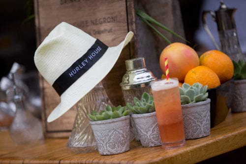 Hurley House - A straw hat with a Hurley House band sits on a wooden table next to a cocktail, succulents, and citrus fruits. A shaker and glass bottles are in the background.