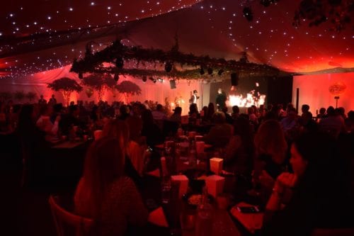 Hurley House - A lively indoor event with a red and white starry-themed ceiling. Attendees sit at tables watching performers on stage. The setting has a festive ambiance with dim lighting and decorative foliage hanging above.