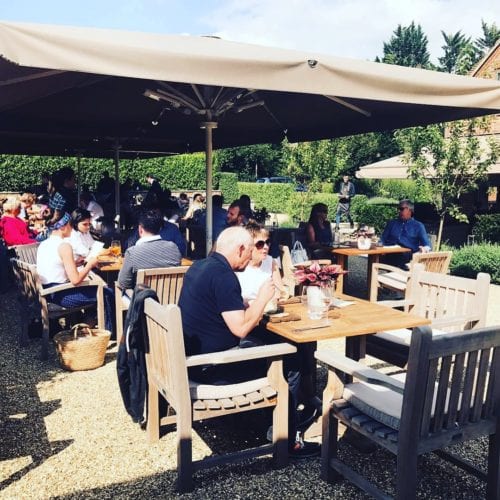 Hurley House - Outdoor restaurant scene with people seated at wooden tables under large umbrellas. Patrons are enjoying meals and drinks on a sunny day, surrounded by greenery and a hedge in the background. Casual and relaxed atmosphere.