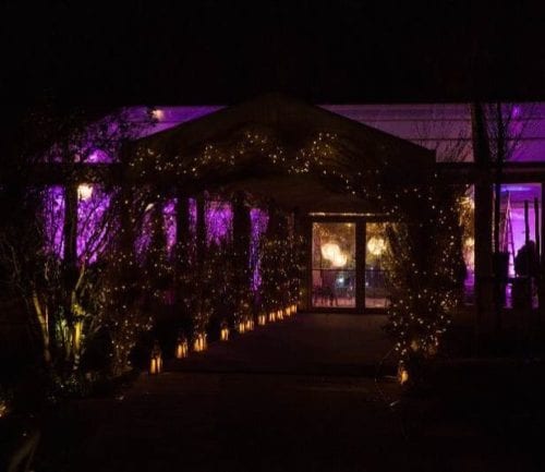 Hurley House - A dimly lit outdoor structure with a canopy decorated with string lights and lanterns, leading to large glass doors. The scene is illuminated with purple lighting, creating a festive and inviting atmosphere at night.