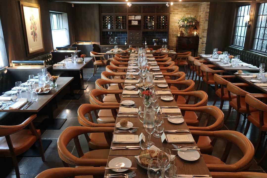 Hurley House - A long, elegantly set dining table is prepared for an event, with neatly arranged plates, glasses, and cutlery. The warm, inviting room has soft lighting, tan leather chairs, and wine bottles displayed on shelves in the background.