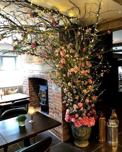 Hurley House - A cozy cafe corner with a wood-burning stove in a brick wall. A large floral arrangement with pink roses and twinkling lights extends over the seating area, which features wooden tables and plush chairs. Soft lighting creates a warm atmosphere.