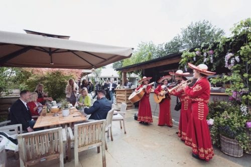 Hurley House - A mariachi band in red outfits performs outdoors for people sitting at wooden tables. The setting includes leafy green plants, umbrellas, and a relaxed atmosphere.