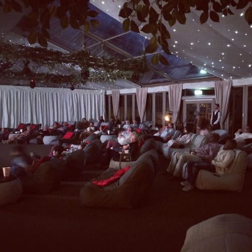 Hurley House - People are seated on large, cushioned recliners in a dimly lit room, watching a projection at the front. The ceiling is decorated with string lights and greenery, creating a cozy ambiance. The audience appears relaxed and engaged.
