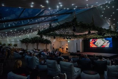 Hurley House - A cozy, dimly lit event space with a large screen showing a colorful image. Attendees sit in plush chairs, surrounded by greenery and string lights on the ceiling, creating a relaxed, outdoor-like atmosphere.