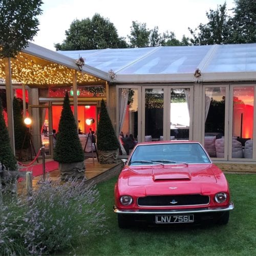 Hurley House - A vintage red sports car parked on grass in front of a clear-roofed event tent with warm lighting. Triangular topiary trees line a red carpet leading into the tent.