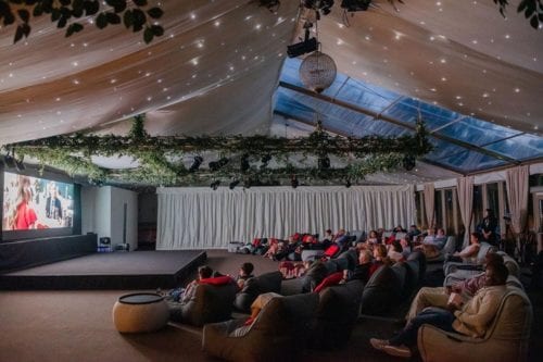 Hurley House - A large group of people sit in a spacious, elegantly decorated room with a glass ceiling. They are watching a movie on a large screen. The ceiling is adorned with greenery and twinkling lights, and there are comfortable bean bags for seating.