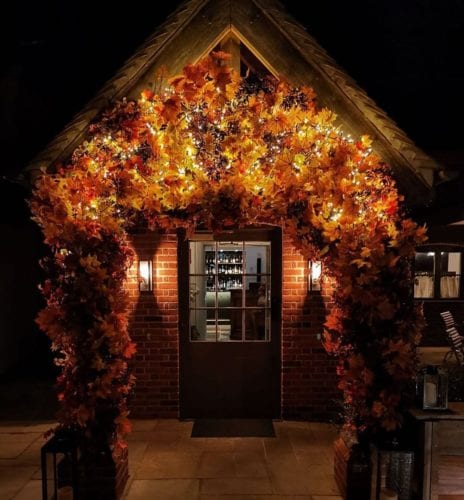 Hurley House - A small brick building with a fall-themed arch made of colorful autumn leaves and twinkling lights at the entrance. Two wall-mounted lanterns illuminate the scene, adding warmth to the cozy, night-time setting.
