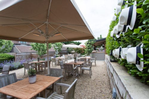 Hurley House - Outdoor patio with wooden tables and chairs under large umbrellas. A wall adorned with white hats on the right adds a decorative touch. Lush greenery surrounds the area, creating a cozy and inviting atmosphere.