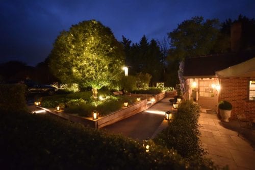Hurley House - A serene evening garden scene with lanterns lining a pathway. A large tree adorned with fairy lights stands illuminated, and a cozy building with warm lights is visible to the right, surrounded by hedges. The sky is a deep blue.