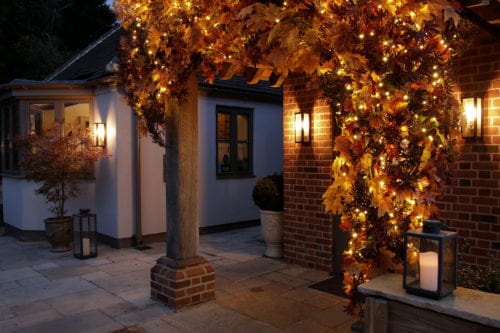 Hurley House - A cozy house entrance decorated with warm string lights and autumn leaves on a brick pillar. Lit lanterns and a small tree in a pot enhance the inviting ambiance. The scene is illuminated, creating a warm and welcoming atmosphere.