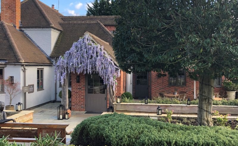Hurley House - A charming brick house with a gabled roof, adorned with blooming wisteria. A neatly trimmed hedge surrounds the garden, and a large tree provides shade. Outdoor wooden benches and lanterns decorate the paved patio.