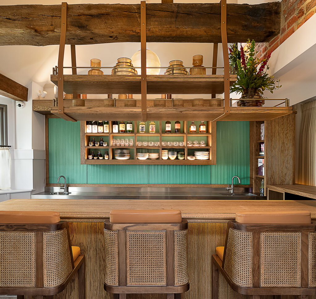 Hurley House - A cozy kitchen with a wooden island, three cane-backed bar stools, green paneling, open shelves displaying dishes and jars, hanging racks with glass containers, exposed beams, and a vase of flowers.