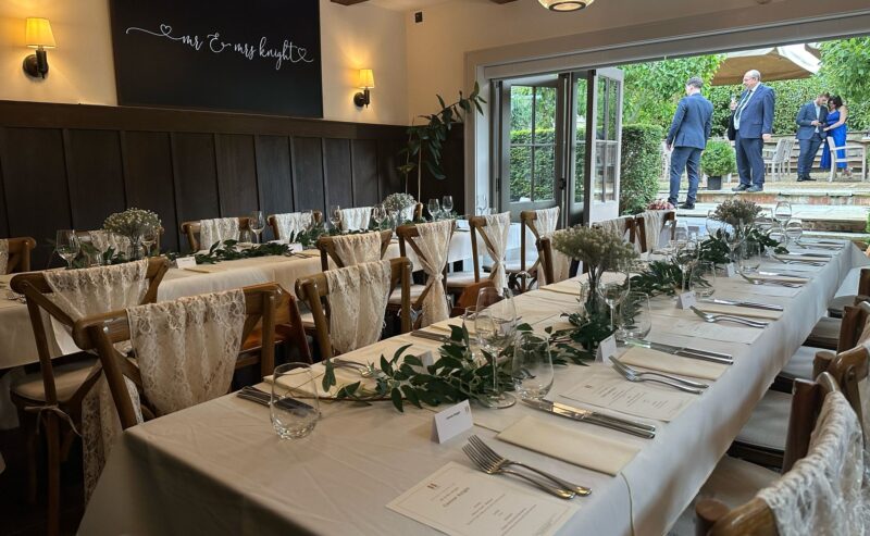 Hurley House - An elegantly set table with white linens and greenery is prepared for a luxury wedding reception in Berkshire. White chairs with lace decorations line the table, beneath a sign reading Mr & Mrs in cursive. Through open glass doors, two people talk outside.