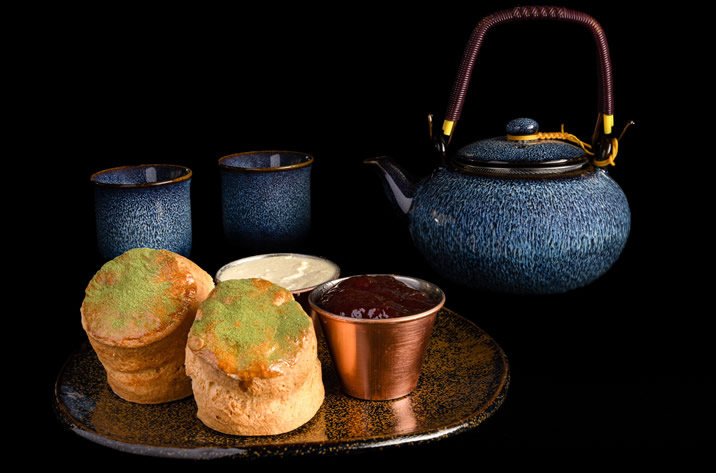 Hurley House - A dark ceramic teapot with two matching cups sits beside a plate holding two scones, a cup of cream, and a cup of red jam, all set against a black background.