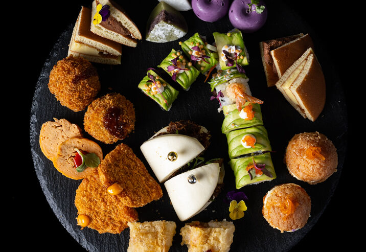 Hurley House - A round black platter with assorted colorful bite-sized appetizers and desserts, including sushi rolls, purple pastries, mini sandwiches, fried bites, steamed buns, and layered cakes, all artfully arranged on a black background.