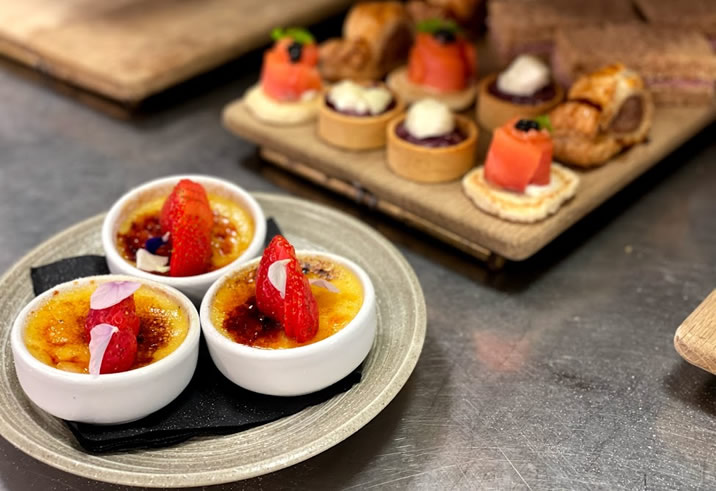 Hurley House - Three small bowls of crème brûlée topped with strawberries and petals sit on a plate in the foreground, while assorted canapés and pastries are displayed on a wooden platter in the background.