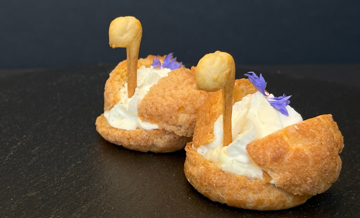 Hurley House - Two cream puffs are decorated to resemble swans with piped cream, pastry necks and heads, and small purple edible flowers, presented on a dark surface against a black background.