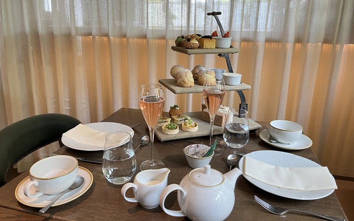 Hurley House - A table set for afternoon tea with white teacups, saucers, a teapot, glasses of pink beverage, and a three-tiered stand holding assorted pastries, bread, and desserts in front of sheer curtains.