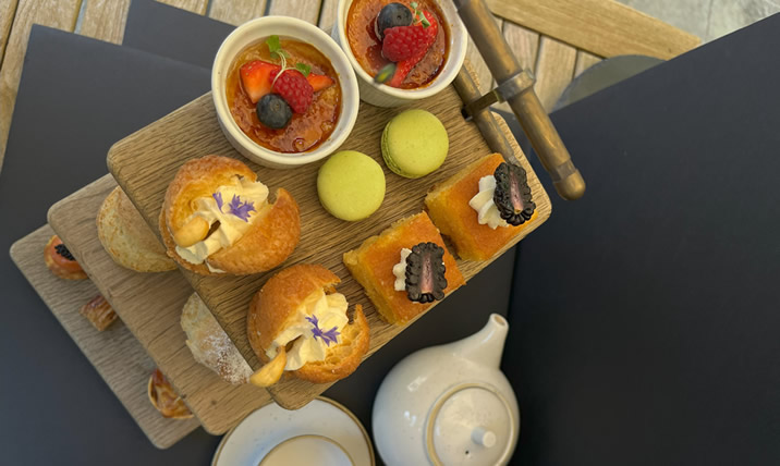 Hurley House - A tiered wooden tray holds assorted pastries, scones, mini cakes topped with berries, and green macarons next to a white teapot and cups on a wooden table.