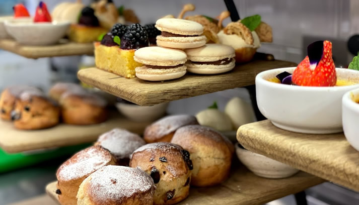 Hurley House - A close-up of wooden trays displaying assorted pastries, including scones dusted with powdered sugar, macarons, fruit-topped cakes, and bowls of dessert garnished with strawberries.