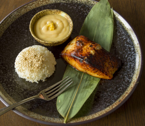 Hurley House - A plate with grilled fish on a green leaf, a serving of white rice topped with sesame seeds, a small bowl of creamy sauce, and a fork, all set on a dark ceramic plate on a wooden table.
