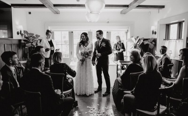 Hurley House - A black and white photo of a wedding ceremony. A bride and groom stand together, smiling, at the front of a room filled with seated guests. Natural light streams in from windows behind them, illuminating rose petals scattered on the floor.
