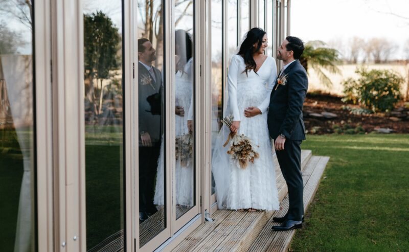 Hurley House - Bride and groom stand on a wooden deck next to large glass doors, which reflect their image. The bride holds a bouquet and wears a long white dress, while the groom is in a gray suit. At this luxury wedding venue in Berkshire, they gaze lovingly amid a backdrop of green grass and trees.