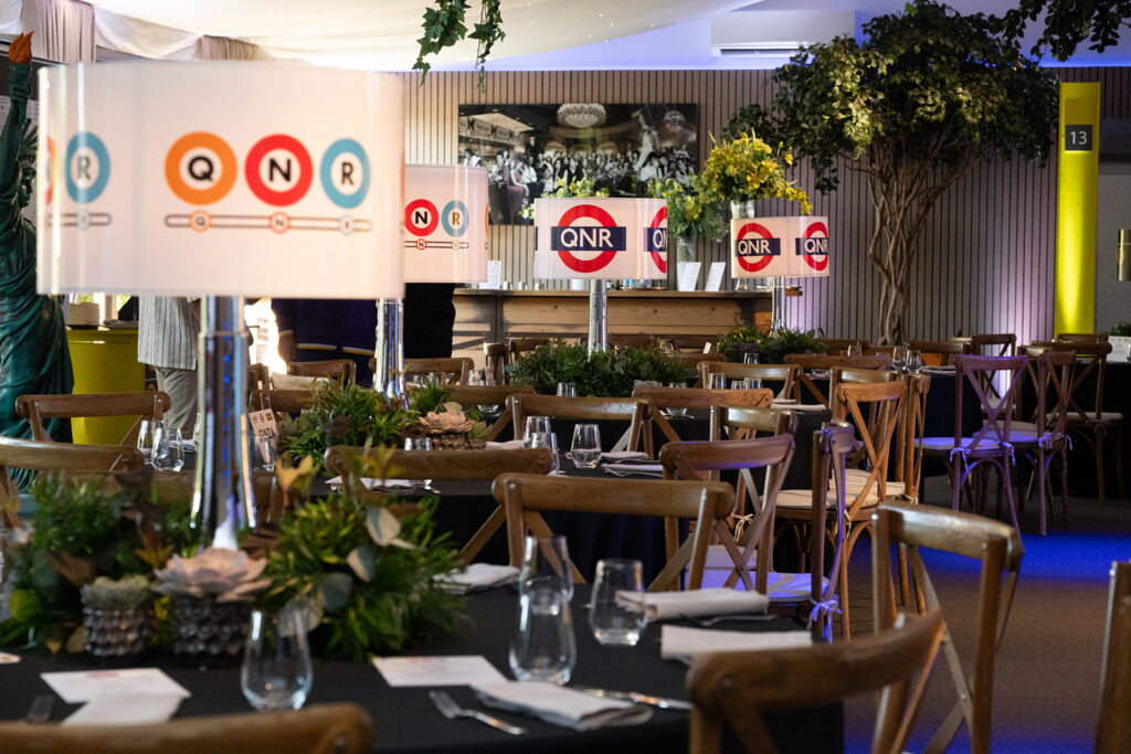 Hurley House - A decorated indoor dining area with round tables, wooden chairs, potted plants, and tall lamps featuring QNR logos. Table settings include glasses and cutlery; greenery and framed photos adorn the space.