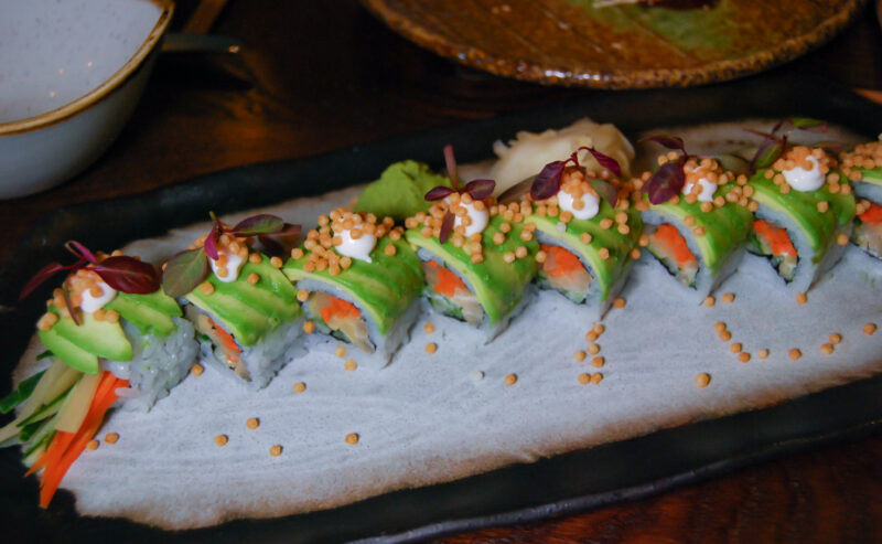 Hurley House - A platter of sushi rolls topped with avocado slices, cream, microgreens, and crunchy golden bits, neatly arranged on a rectangular black plate with wasabi and pickled ginger on the side.