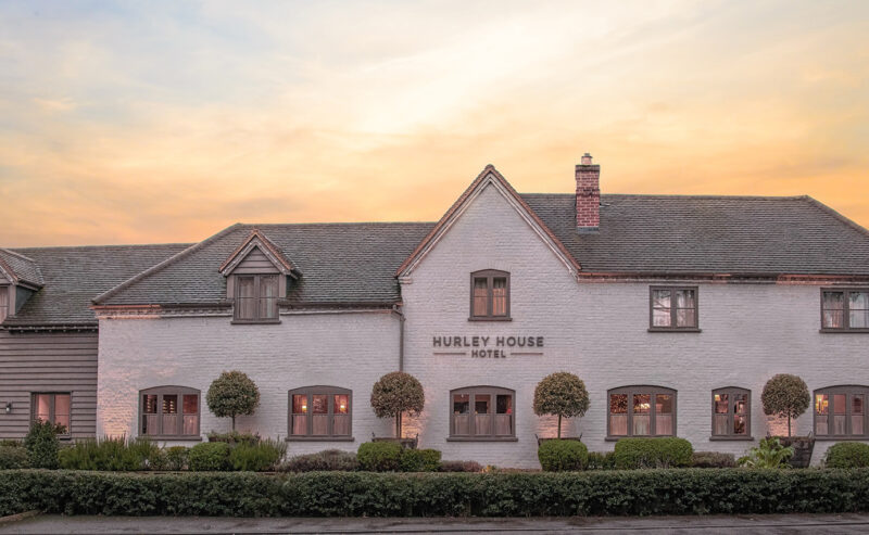 Hurley House - A charming hotel with a rustic exterior and neatly trimmed bushes, set against a peaceful sunset sky. The building is painted white with gray accents and has a sign that reads Hurley House Hotel.