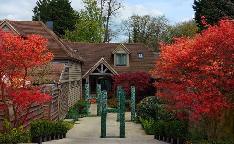 Hurley House - A house with steep gabled roofs is framed by vibrant red-orange trees and green shrubs along a paved pathway under a partly cloudy sky.