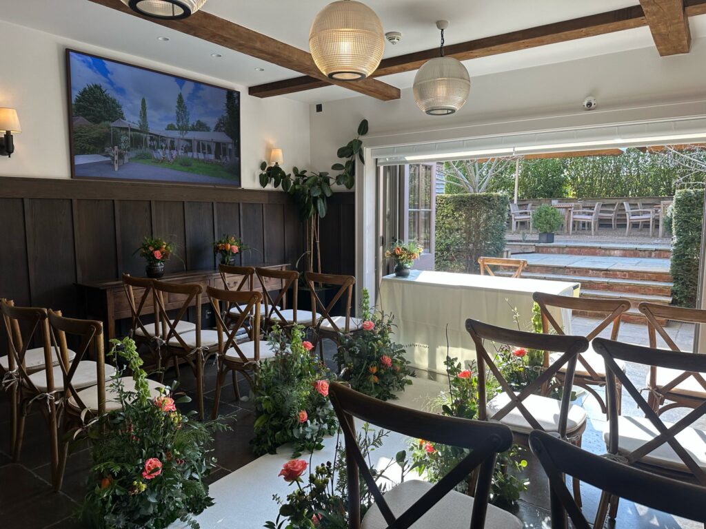Hurley House - A small, elegantly decorated room in a luxury wedding venue in Berkshire is set up for a ceremony. Wooden chairs are arranged in rows amidst lush floral arrangements. Large windows flood the space with natural light, and a photo adorns the wall. A table is gracefully positioned at the front.
