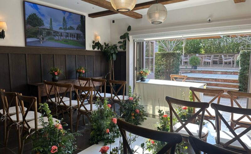 Hurley House - A small, elegantly decorated room in a luxury wedding venue in Berkshire is set up for a ceremony. Wooden chairs are arranged in rows amidst lush floral arrangements. Large windows flood the space with natural light, and a photo adorns the wall. A table is gracefully positioned at the front.