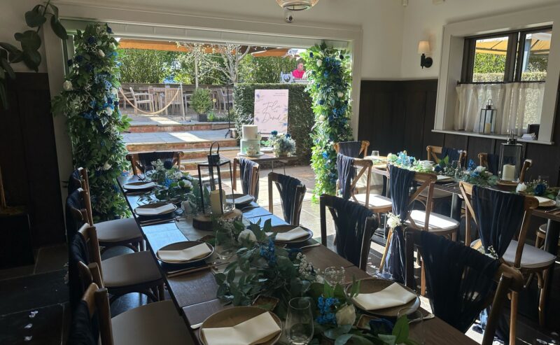 Hurley House - Elegant dining area with wooden tables and chairs, decorated with green foliage and blue accents. Outdoor view through an archway adorned with greenery, perfect for a luxury wedding venue in Berkshire. Bright, airy ambiance with paper lanterns hanging from the ceiling.