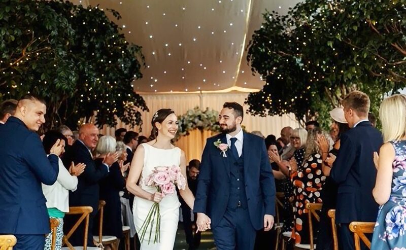 Hurley House - A couple walks down the aisle, holding hands. The bride wears a white dress and holds a bouquet, while the groom wears a dark suit. Guests on either side stand and applaud. The venue is decorated with trees and twinkling lights.