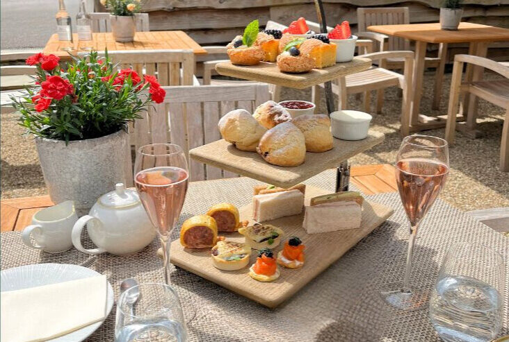 Hurley House - An outdoor table set for afternoon tea with sandwiches, pastries, and scones on wooden trays, glasses of rosé, a teapot, plates, and a vase of red flowers. Wooden benches and greenery are in the background.
