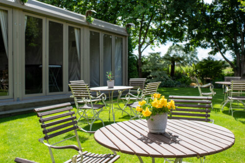 Hurley House - Round wooden tables with metal chairs are arranged on a sunny green lawn. A potted plant with yellow flowers sits on a table in the foreground. Large windows and trees provide shade in the background.