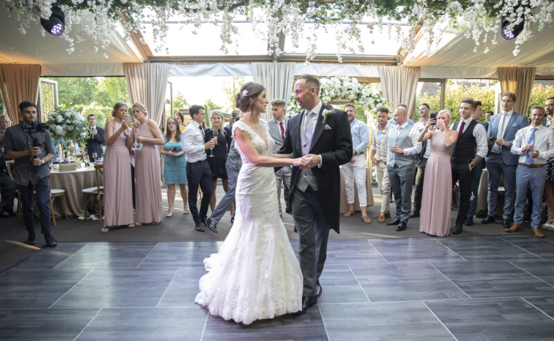 Hurley House - In a luxury wedding venue in Berkshire, the bride and groom share their first dance under soft lighting, surrounded by guests. She dazzles in a lace wedding dress, while he looks dapper in his suit. The room is adorned with elegant floral arrangements, creating an enchanting atmosphere.