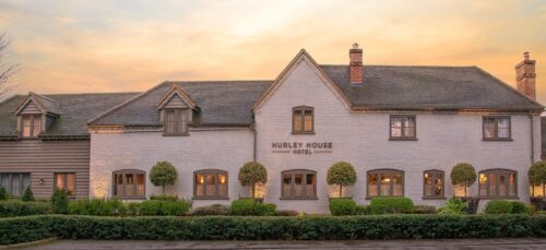 Hurley House - A large, rustic-style building with a sign reading Hurley House Hotel on the front, this luxury wedding venue in Berkshire features grey bricks, a slate roof, and several windows. The entrance is framed by neatly trimmed bushes under a warm, glowing sunset sky.