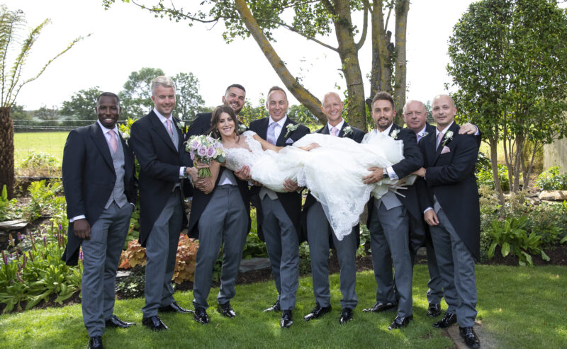 Hurley House - A bride in a lace wedding dress is playfully held horizontally by groomsmen in matching suits with pink ties, all smiling against the backdrop of a luxury wedding venue in Berkshire, surrounded by lush gardens and a sunny sky.