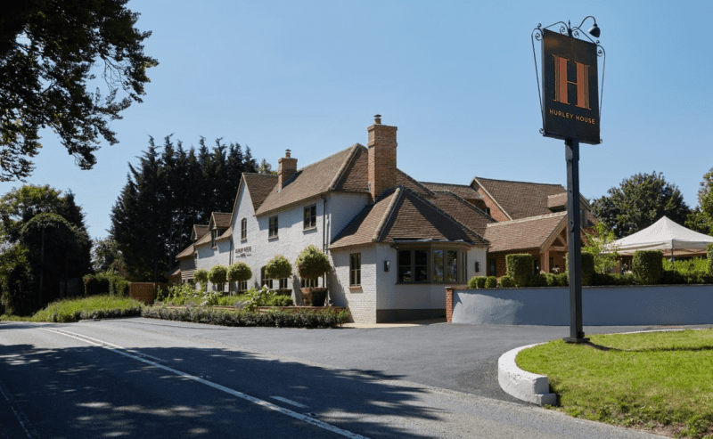 Hurley House - A well-maintained, two-story brick building with a pitched roof, surrounded by greenery on a sunny day. A large sign in front reads Hurley House. Theres a clear, blue sky above the building nestled by trees.