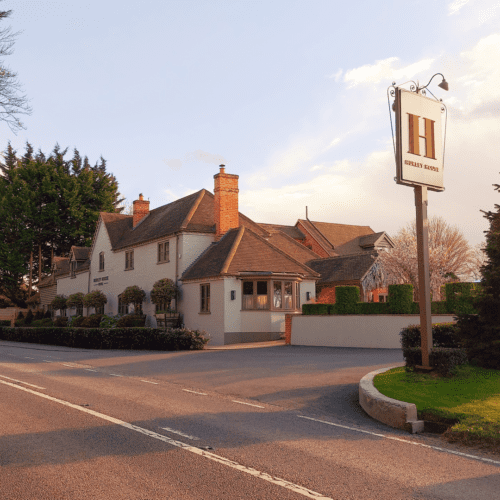 Hurley House - A quaint country hotel with a beige facade and brown roof sits beside a spacious parking area. The sign reads Hurley House Hotel. Tall trees border the property, and the sky is clear with soft sunlight illuminating the scene.