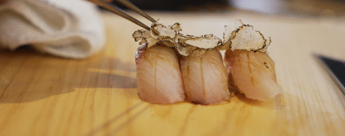 Hurley House - Three pieces of sushi topped with thinly sliced truffle are arranged on a wooden surface, with a hand using chopsticks to pick up one piece.