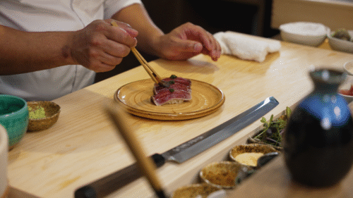 Hurley House - A chef uses chopsticks to plate slices of raw fish on a ceramic plate at a wooden counter, surrounded by bowls of ingredients and a knife.