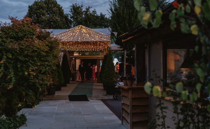 Hurley House - A warmly lit event tent with string lights is nestled among greenery at dusk. People are visible inside the tent, and the scene has a cozy, inviting atmosphere with trees and plants framing the walkway.