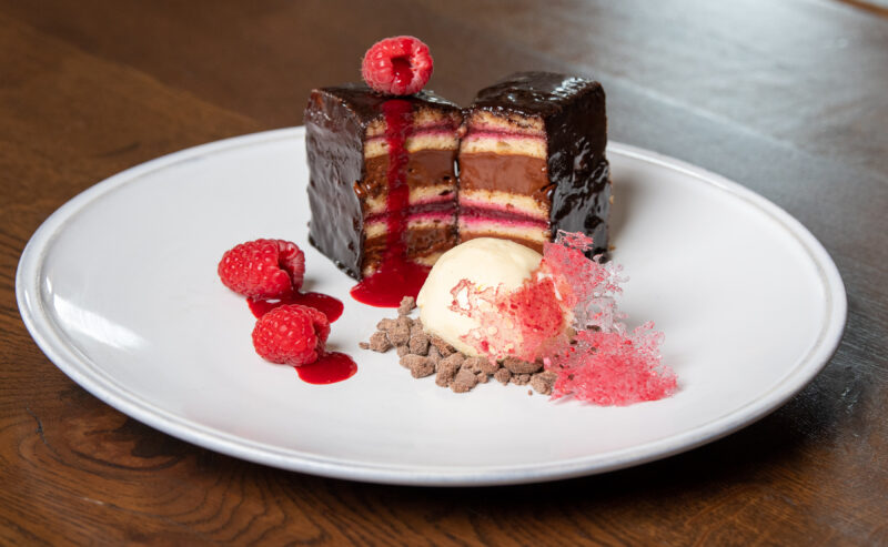 Hurley House - A plated dessert featuring layered chocolate-covered cake with fruit filling, garnished with raspberries, a scoop of vanilla ice cream, chocolate crumbles, and pink sugar tuile on a white plate.