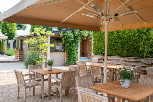 Hurley House - Outdoor patio area with wooden tables and chairs under large beige umbrellas, surrounded by greenery and potted plants, with a wooden building in the background. The setting appears calm and inviting.