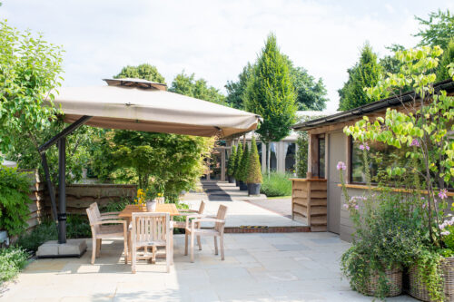 Hurley House - A sunny patio with a wooden table and four chairs under a large beige umbrella, surrounded by green plants and trees, near a house with large windows and potted plants.